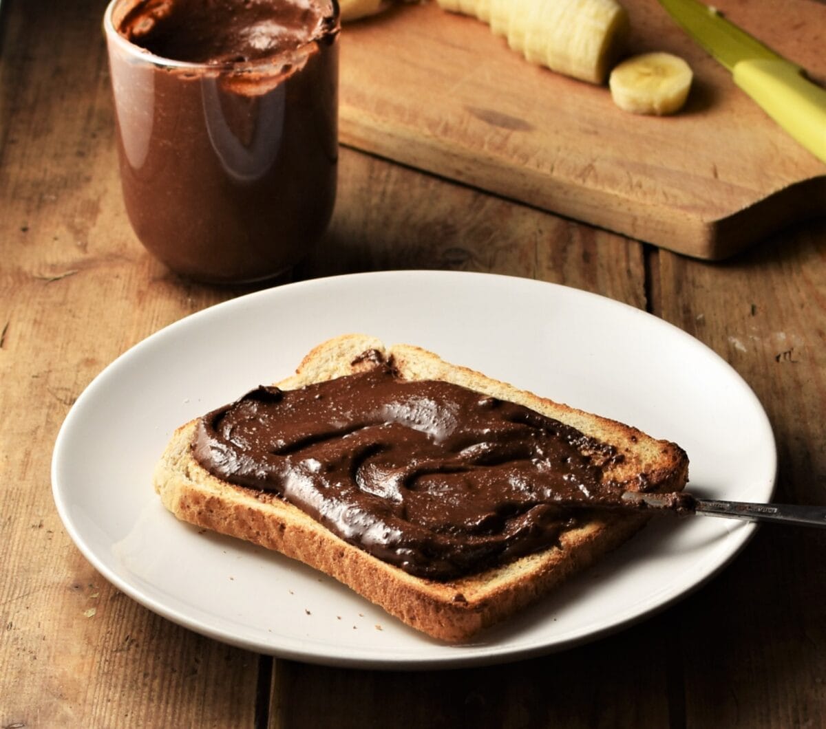 Side view of toast with chocolate hazelnut spread and knife on white plate, with spread in cup and cutting board in background.