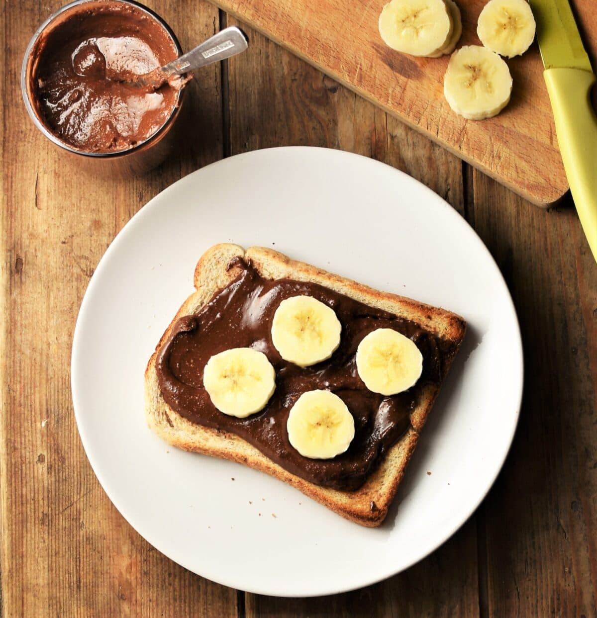 Top down view of hazelnut cacao spread on toast with sliced banana on white plate, with spread in cup and banana in background.