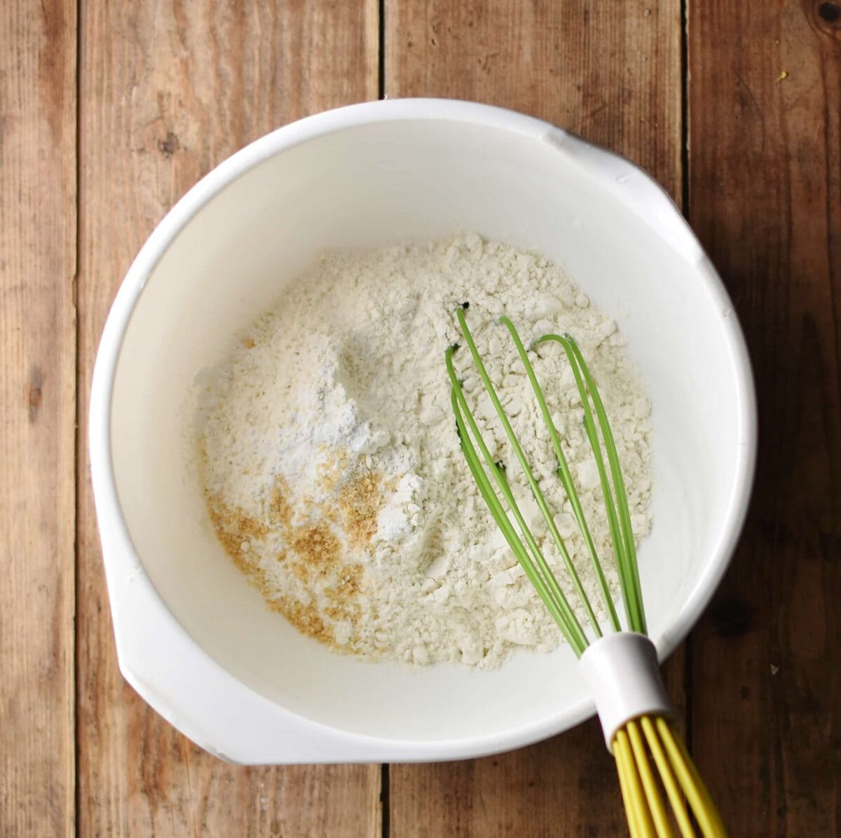 Flour mixture inside large white bowl with green whisk.