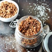 Top down view of chocolate granola in open jar with spoon to right and sea salt on top of dark shiny surface.