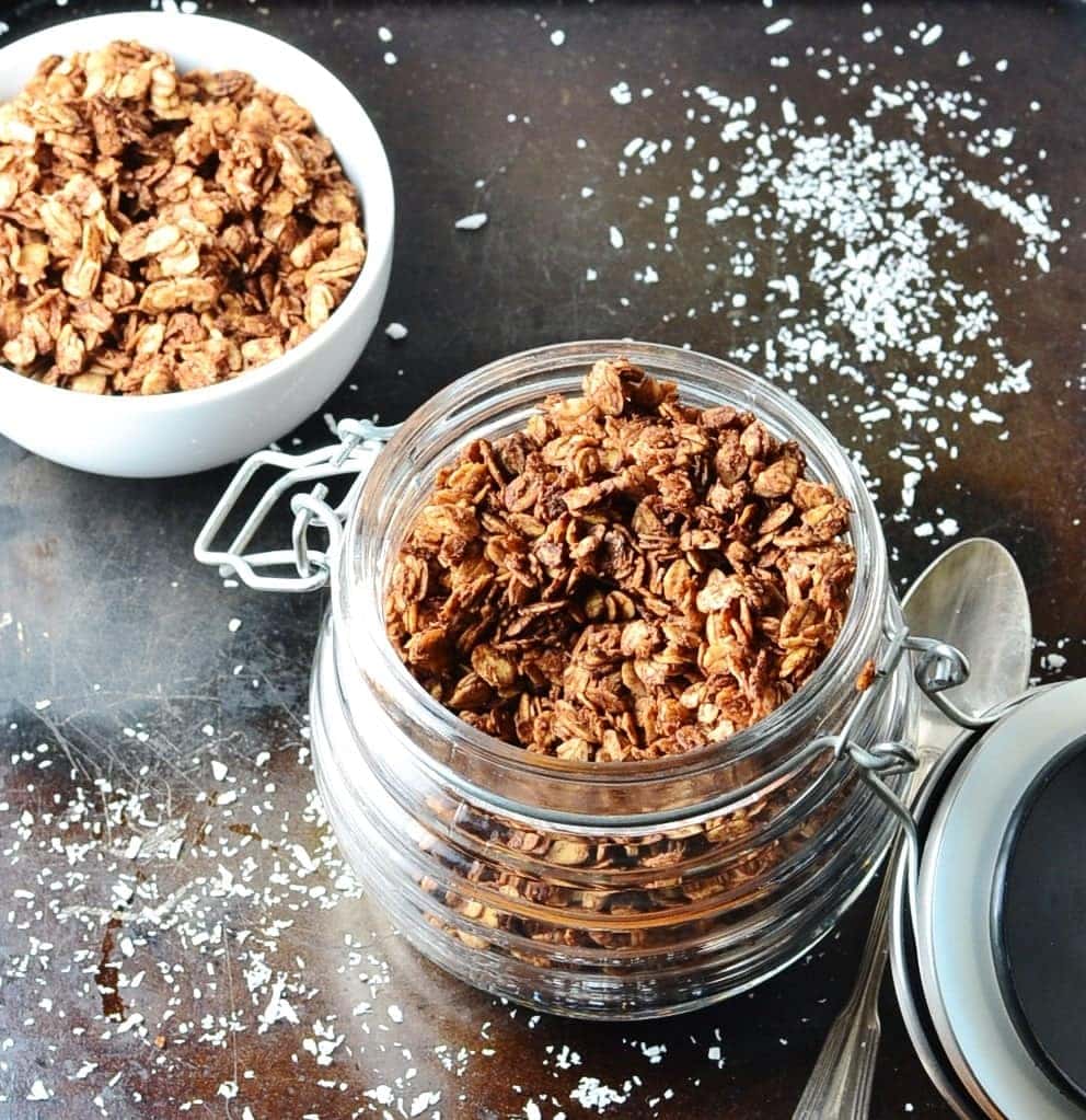 Top down view of chocolate granola in open jar with spoon to right and sea salt on top of dark shiny surface.