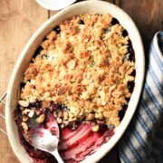 Top down view of cherry crumble in white oval dish with spoon, blue cloth and yogurt in background.