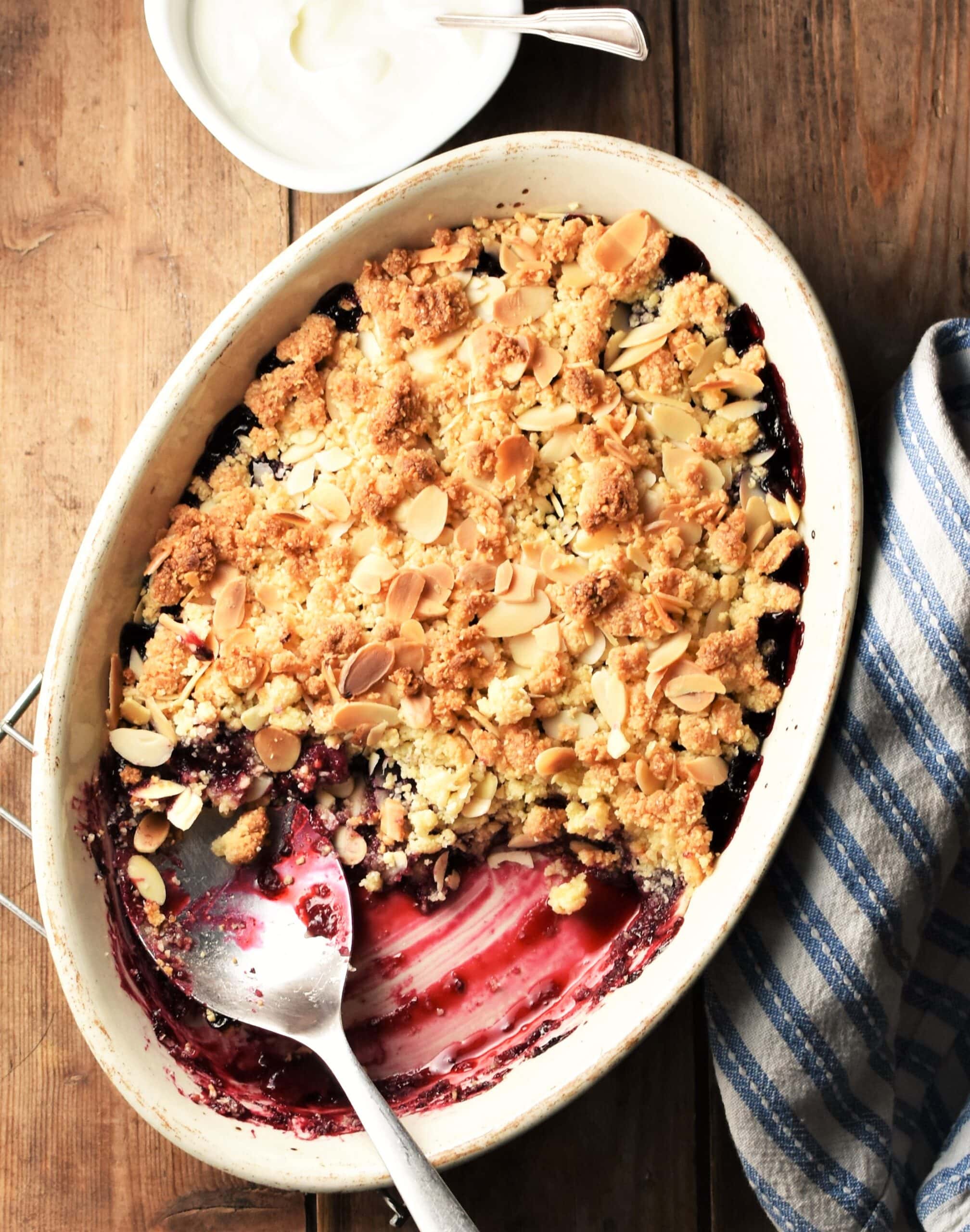 Top down view of cherry crumble in white oval dish with spoon, blue cloth and yogurt in background.
