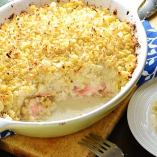 Healthy fish pie with cauliflower topping in white oval casserole dish on wooden board with fork and partial view of white plate with pie.