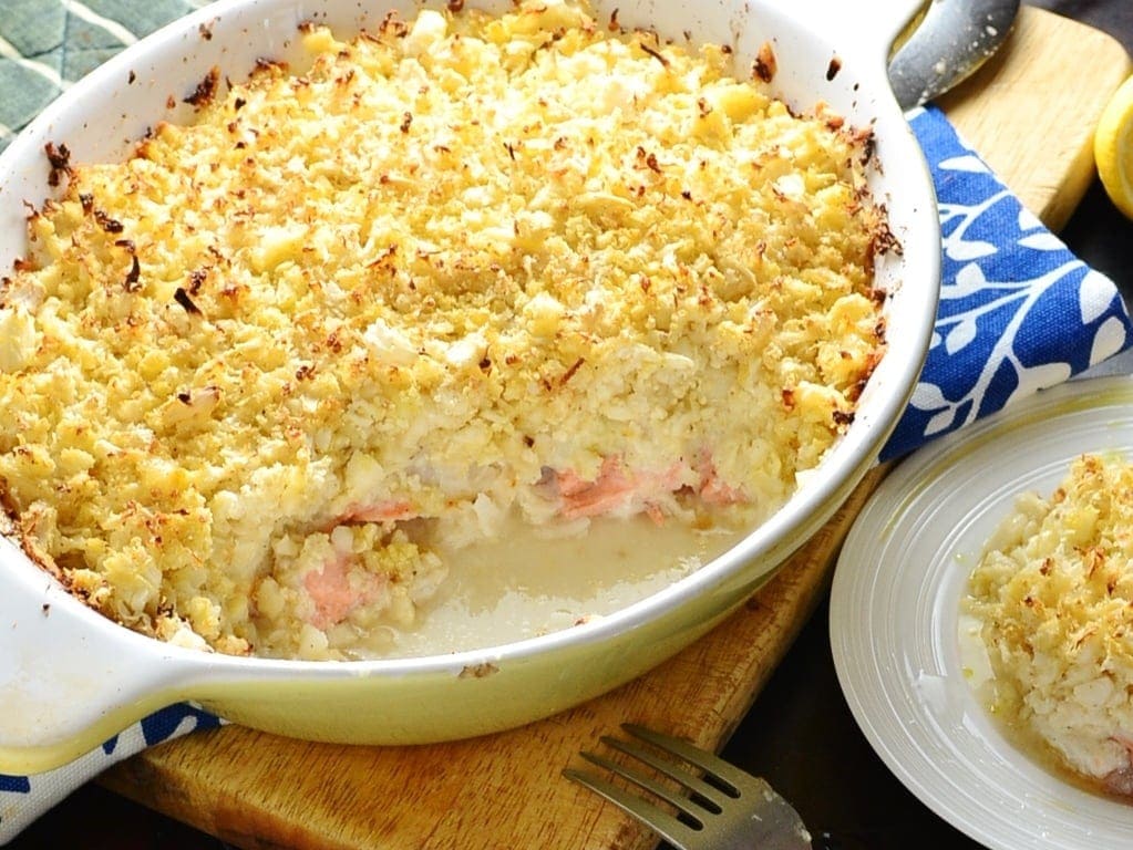Healthy fish pie with cauliflower topping in white oval casserole dish on wooden board with fork and partial view of white plate with pie.