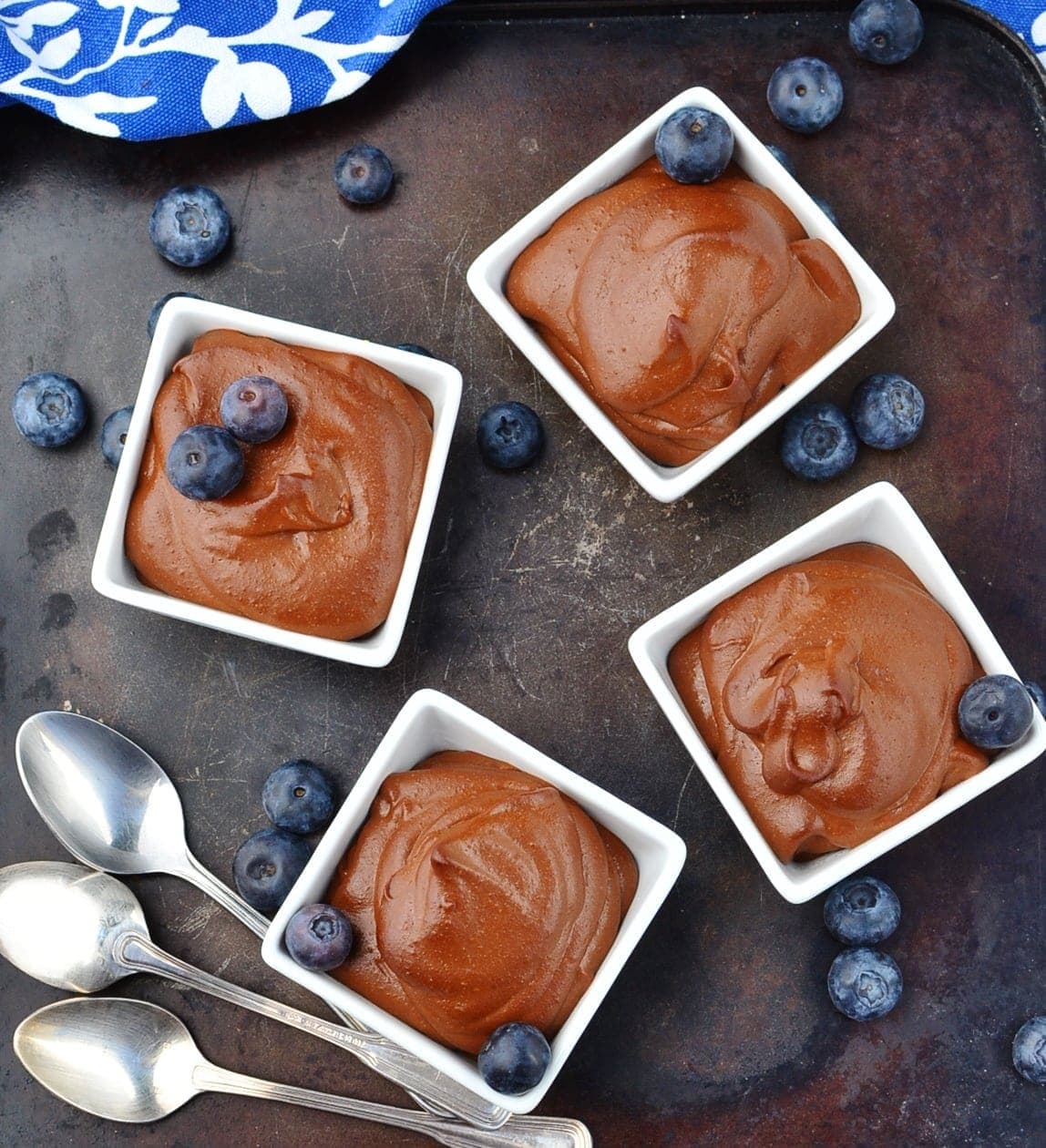 Top down view of chocolate pudding in 4 white square dishes topped with blueberries on top of dark surface with 3 spoons, blueberries and blue-and-white cloth at top.