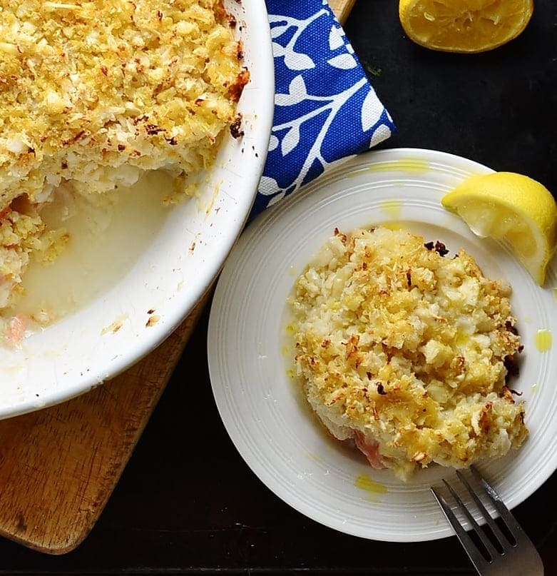 Top down view of fish pie with cauliflower topping on top of white plate with lemon wedge and fork and inside white casserole dish.