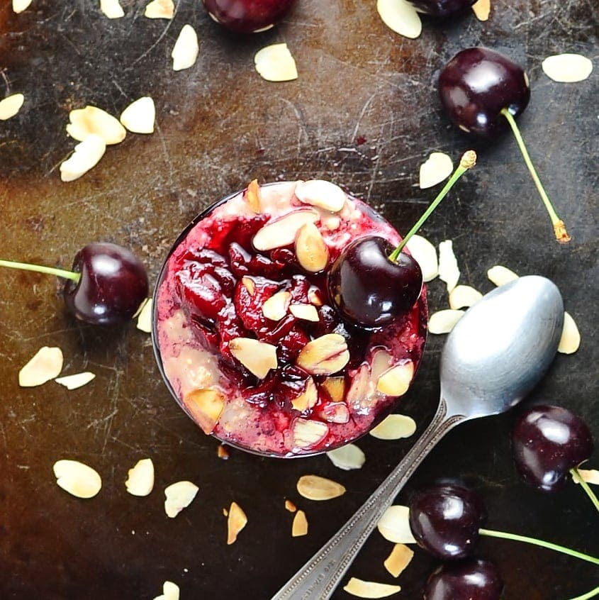 Top down view of cherry overnight oats with spoon, almond flakes and cherries on top of dark tray.