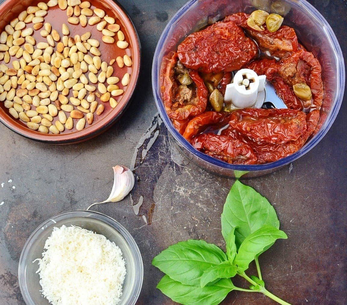 Top down view of sun dried tomatoes in blender, pine nuts, grated cheese in small dish and basil leaves on top of dark surface.