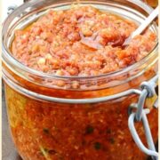 Side view of sun dried tomato pesto in small open jar with spoon, with sun dried tomatoes in small dish and green cloth in background.