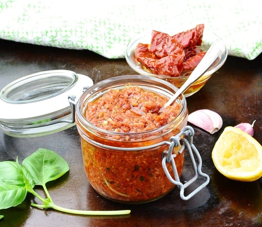 Side view of sun dried tomato pesto in open jar with basil leaves, spoon, lemon, garlic and sun dried tomatoes in background.