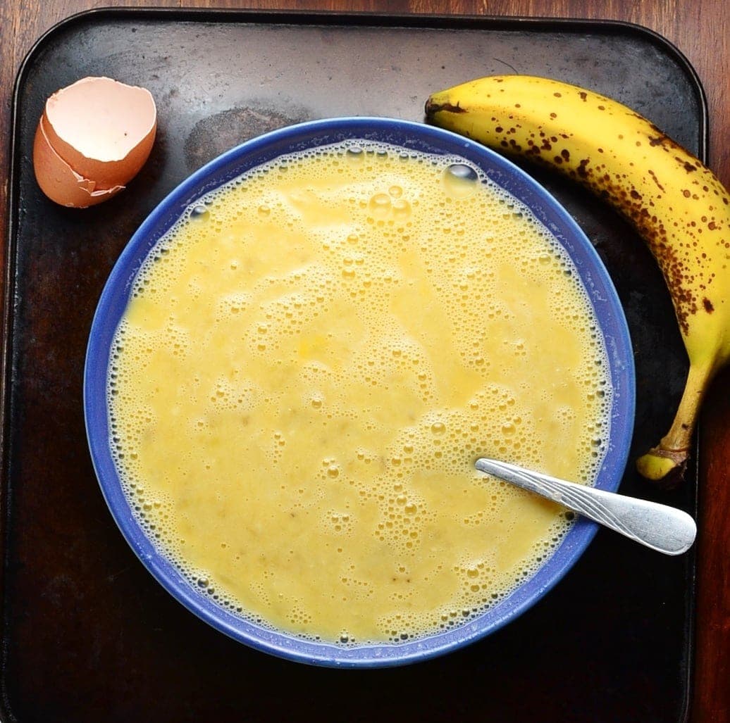 Egg mixture with spoon in blue dish with egg shell in top left and ripe banana in top right on oven tray.