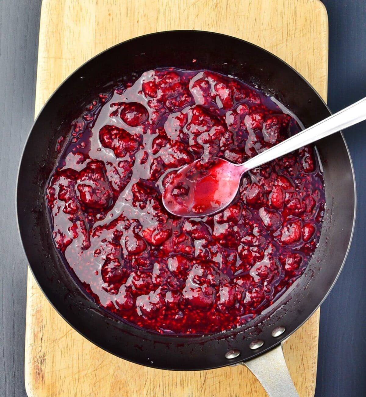 Top down view of cooked raspberries with spoon in skillet on top of wooden board.