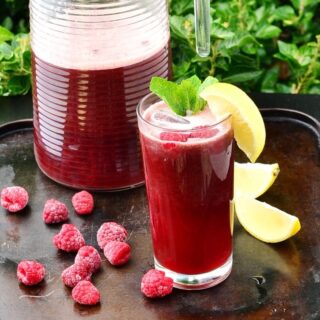 Side view of rooibos iced tea with lemon wedge and mint leaves in tall glass, with frozen raspberries and lemon wedges on top of brown tray, and jug of iced tea in background.