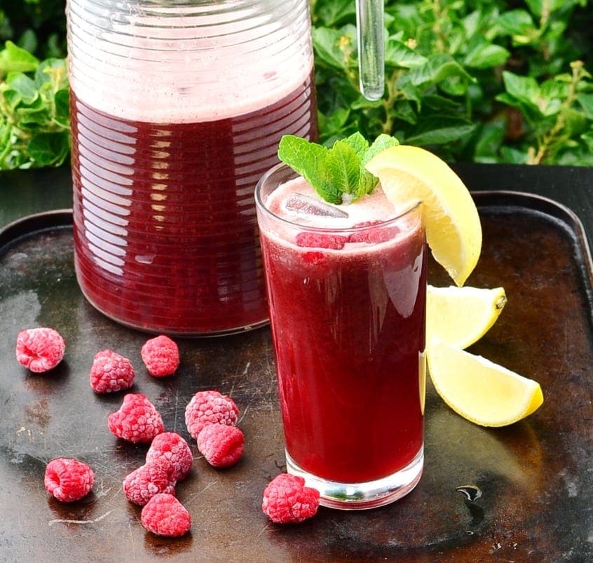 Side view of rooibos iced tea with lemon wedge and mint leaves in tall glass, with frozen raspberries and lemon wedges on top of brown tray, and jug of iced tea in background.