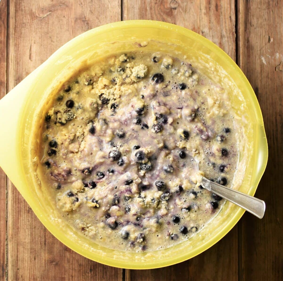 Lumpy blueberry muffin batter in large yellow bowl with spoon.