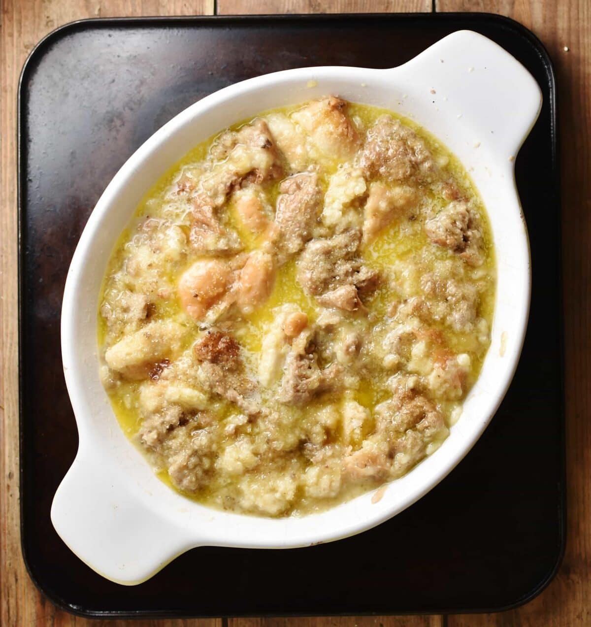 Top down view of torn bread pieces with egg mixture and melted butter inside white oval dish.