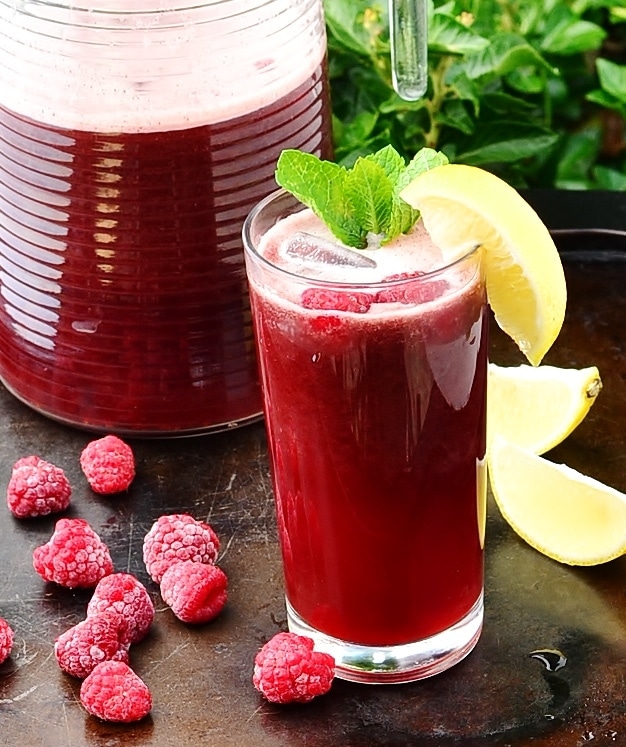 Side view of rooibos iced tea in tall glass with mint leaves, tea in juglemon wedges and raspberries in background.