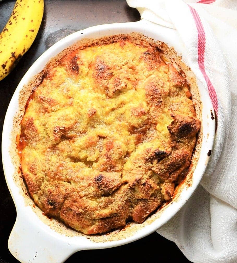 Top down view of banana french toast bake in white oval dish with ripe bananain top left corner and white cloth with red stripe to right.