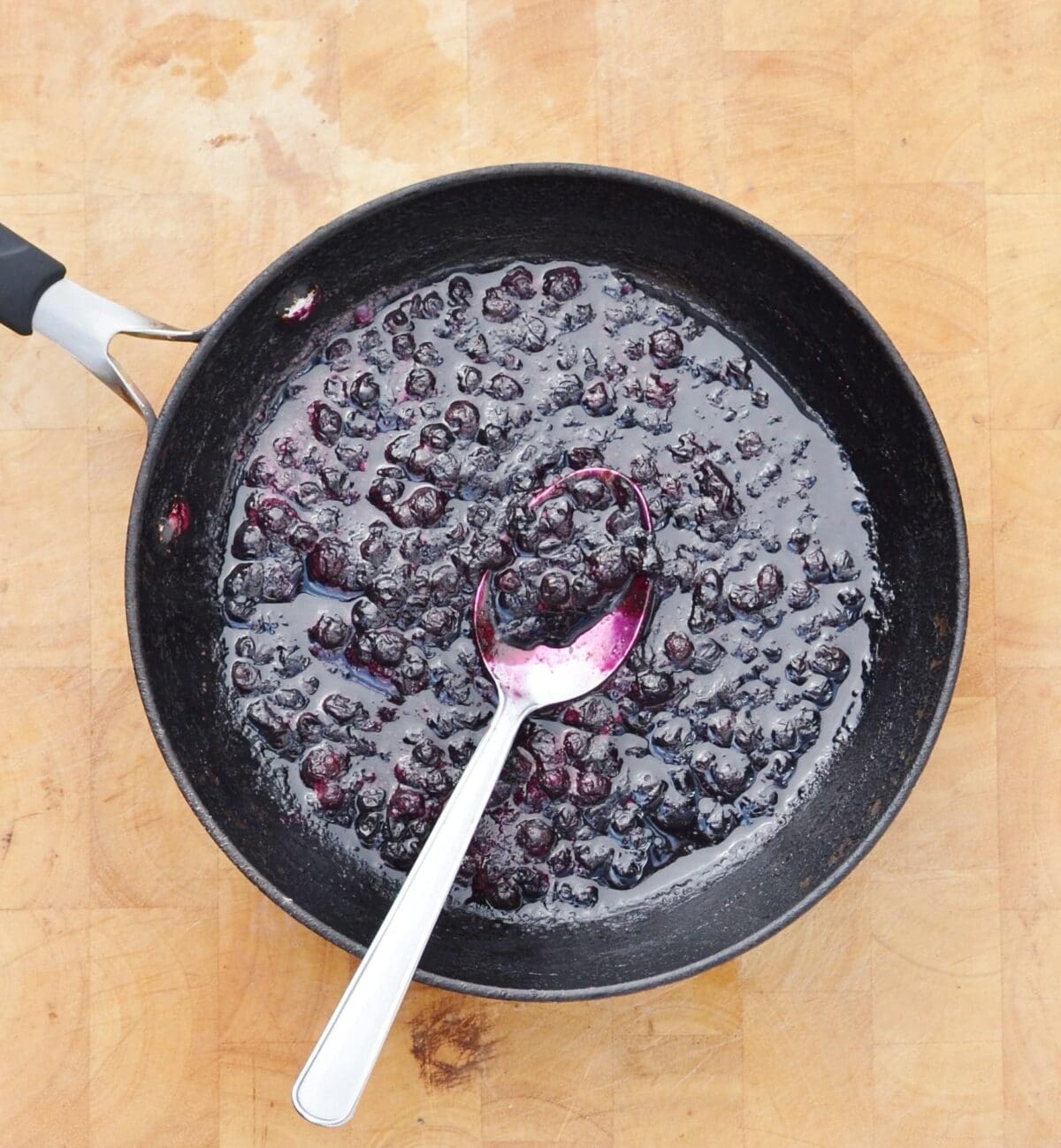 Cooked blueberries with juice in skillet with spoon on light wooden board.