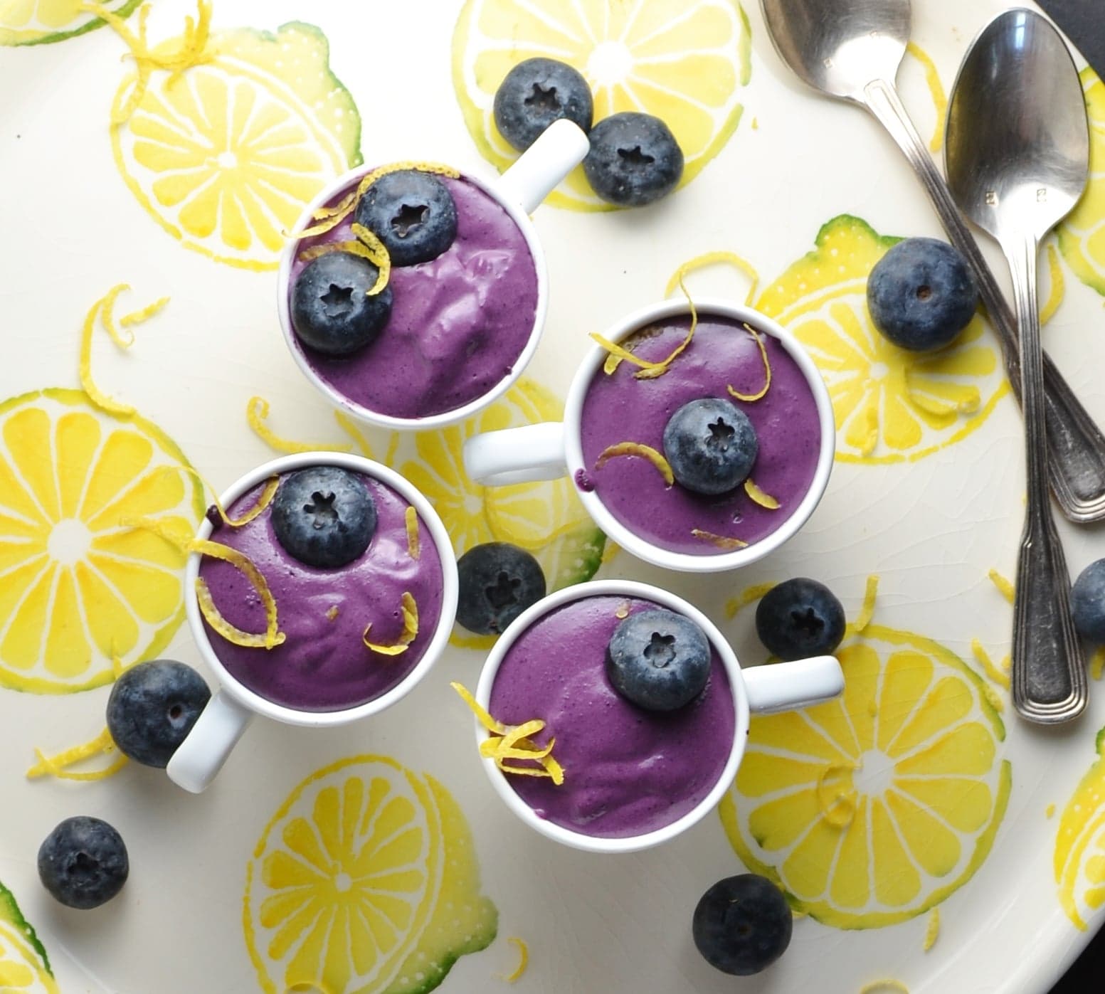 Top down view of blueberry dessert in 4 white cups with garnish of blueberries and lemon zest, with 2 spoons on white plate with lemon pattern.