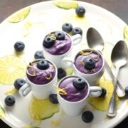 Blueberry cream cheese dessert in 4 white cups with garnish of blueberries and lemon zest, with 2 spoons on white plate with lemon pattern.