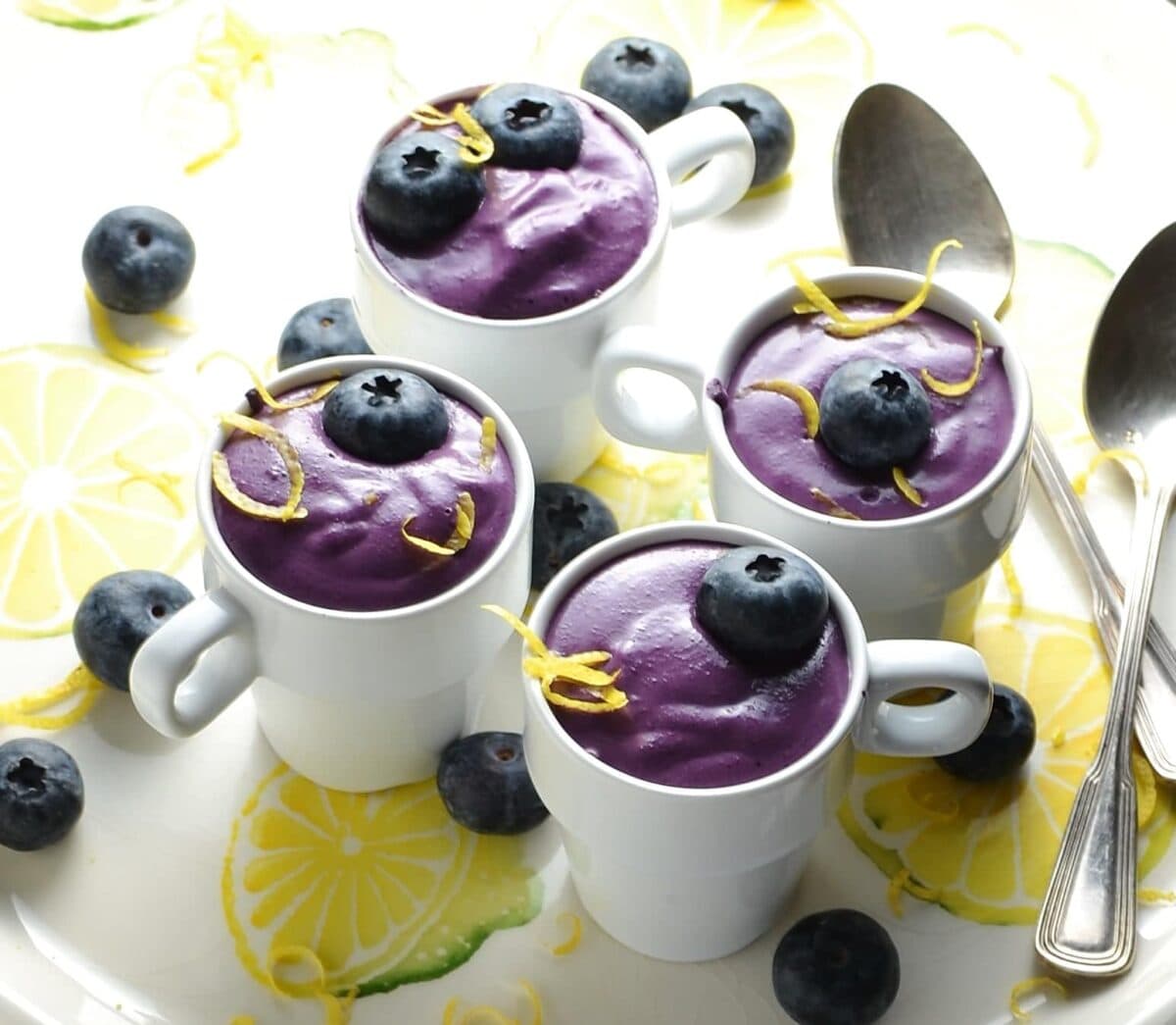 Close-up view of blueberry dessert with fresh blueberries and lemon zest inside small white cups on top of white plate with lemon pattern.