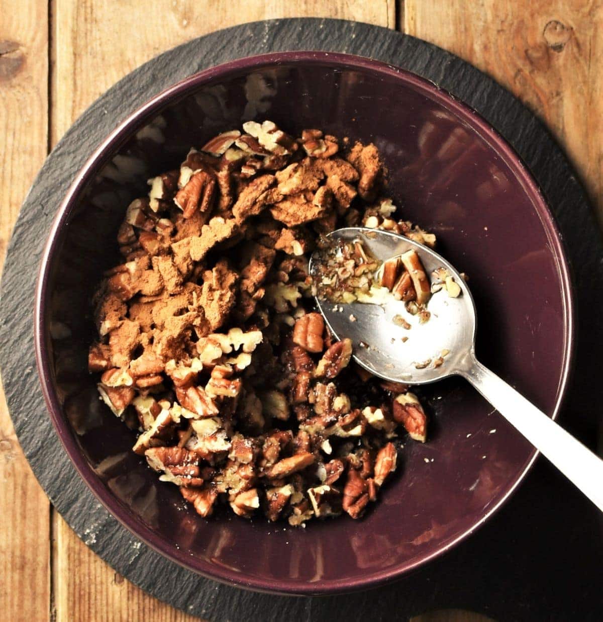 Chopped pecans with cinnamon and spoon in purple bowl.