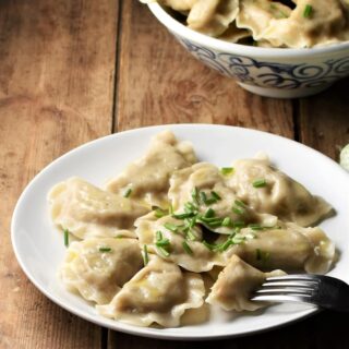 Side view of pierogi on top of plate with pierogi in bowl in background.