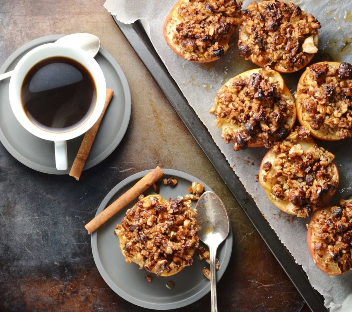 Top down view of apple halves with browned pecans on top of baking sheet and grey saucer, with coffee in white cup.