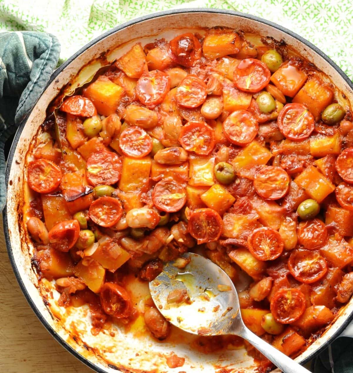 Top down view of butternut squash casserole in large shallow white dish with spoon and green cloth at top.