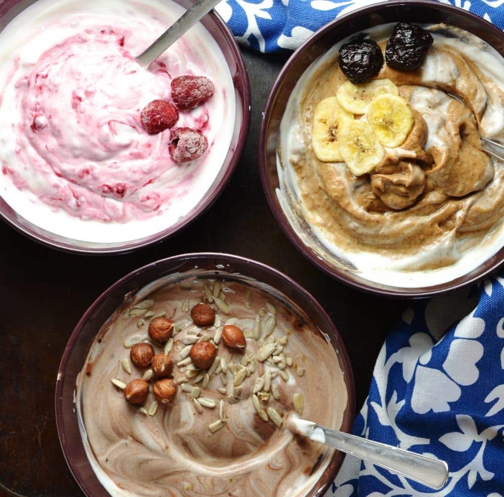 Mocha, raspberry and prune yogurts in separate bowl with spoons, nuts and fruit on top and blue-and-white cloth in background.