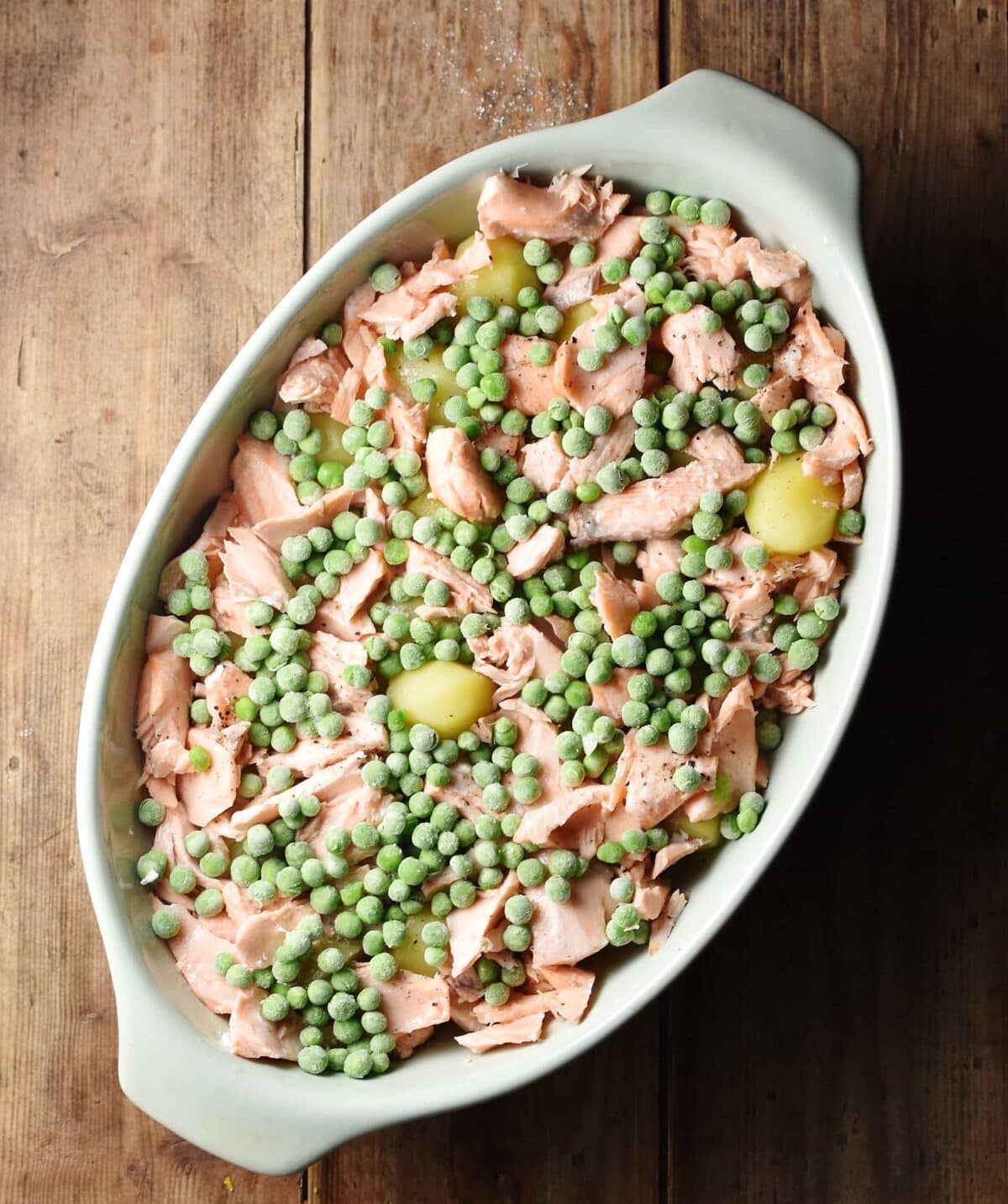 Frozen peas and shredded cooked salmon on top of potatoes in oval, white casserole dish.