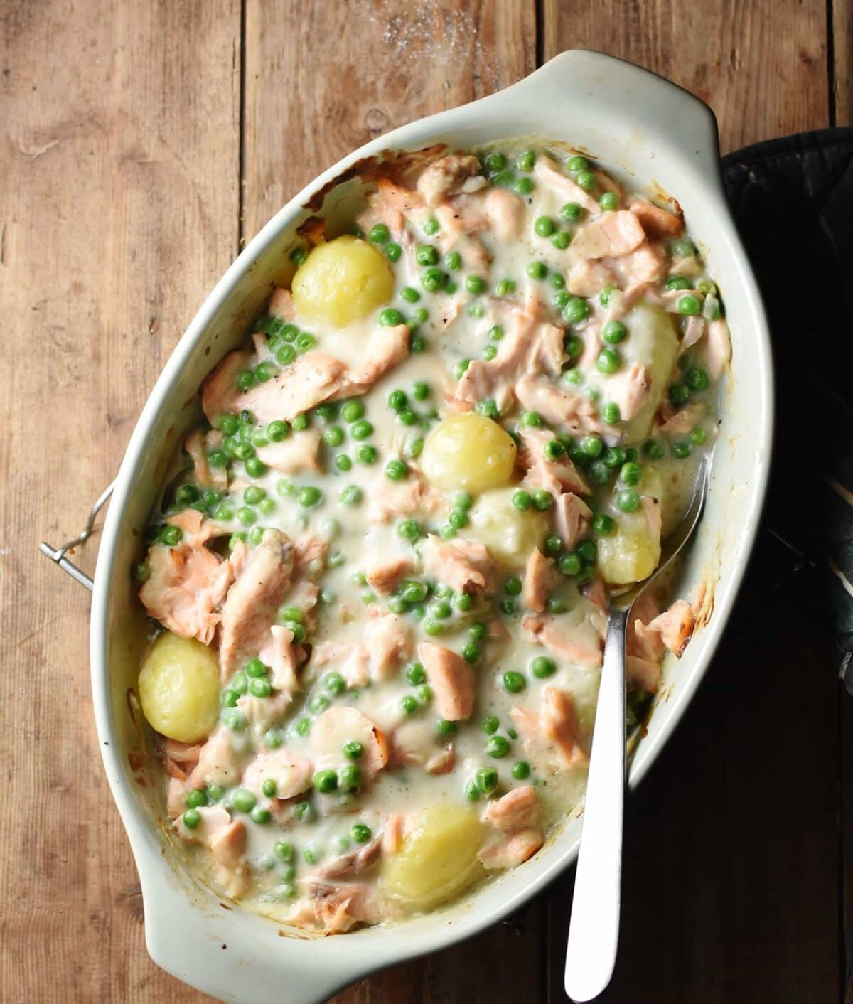 Creamy salmon, potato and peas bake in white, oval casserole dish with spoon.