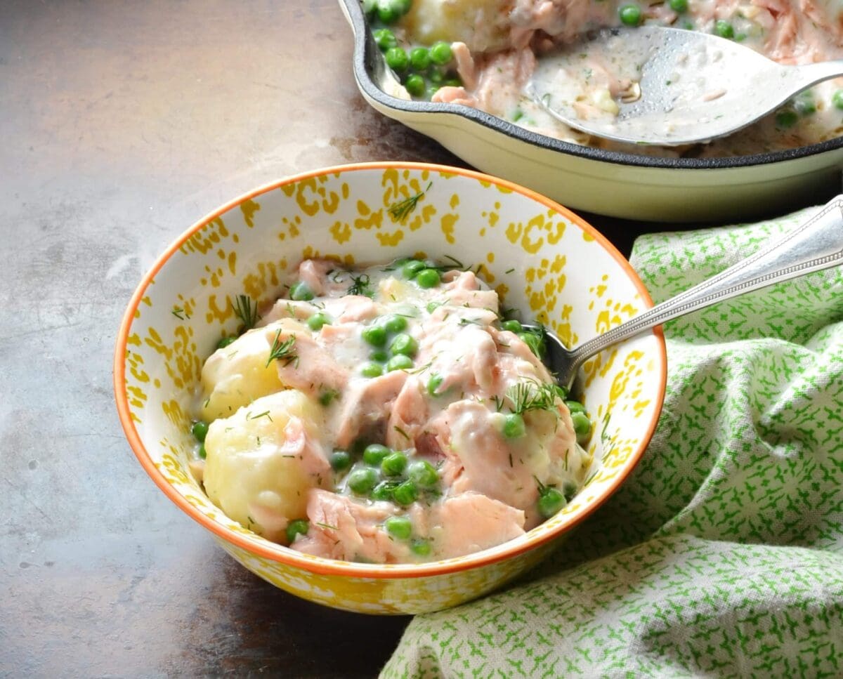 Salmon and potato bake in white-and-yellow bowl with spoon and green cloth and white casserole dish and spoon in background.