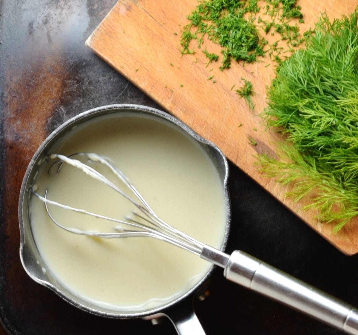 White sauce in black saucepan with whisk and dill on wooden cutting board.