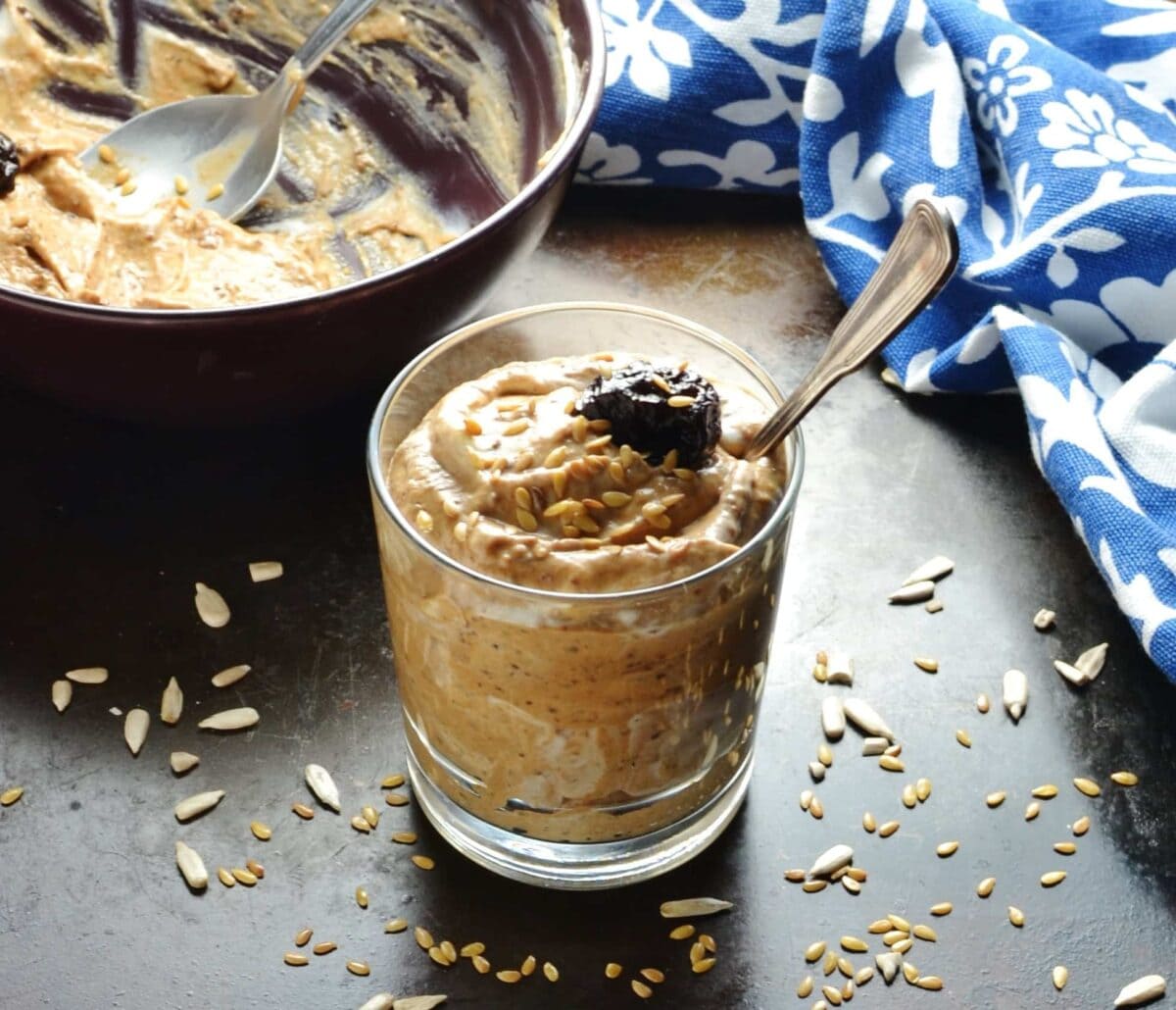 Flavoured prune yogurt in glass with spoon, bowl and blue-and-white cloth in background.