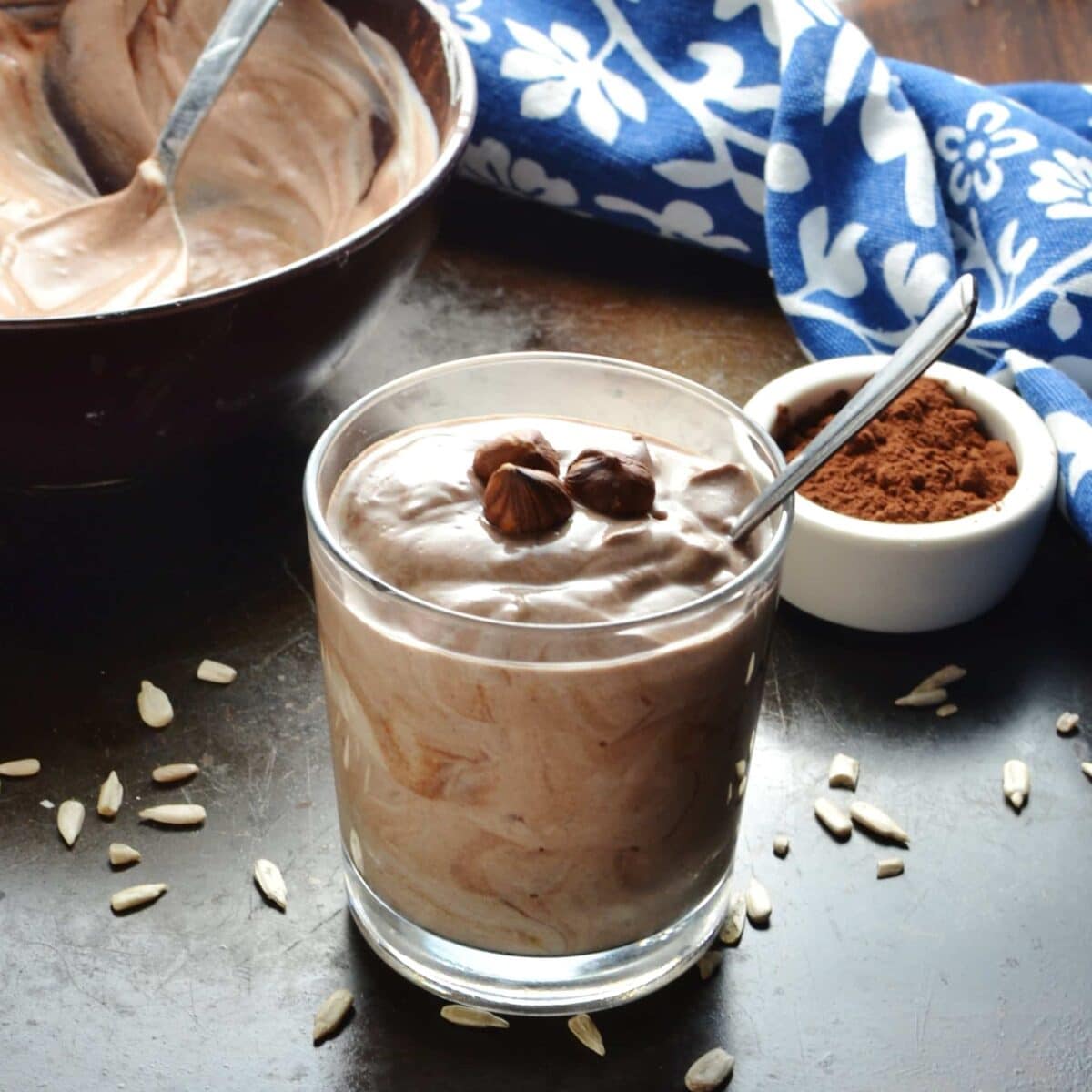 Flavoured mocha yogurt in glass with spoon, purple bowl, white dish with cocoa and blue-and-white cloth in background.