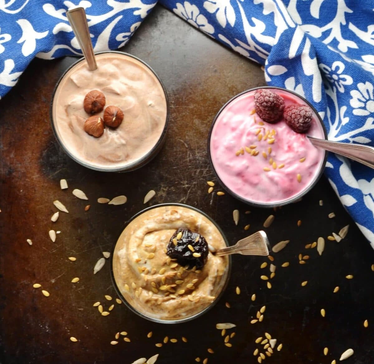 Flavoured mocha, raspberry and prune yogurt in bowls with spoons and blue-and-white cloth on brown surface.