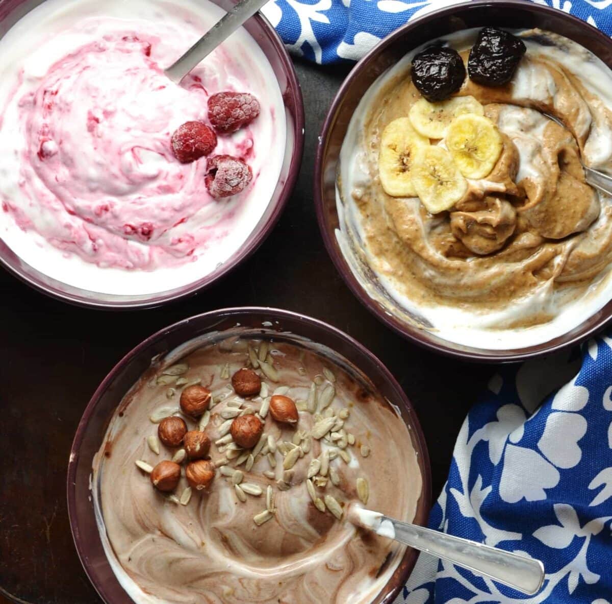 Flavoured mocha, raspberry, prune yogurt in purple bowls with spoons and blue-and-white clothe on dark surface.