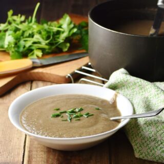 Mushroom potato soup in white bowl with spoon, green cloth, herbs, knife and large pot in background.