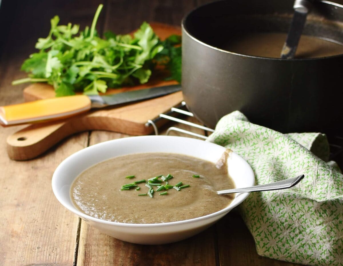 Creamy mushroom soup with chives in white bowl with spoon, green cloth, herbs, knife and large pot in background.