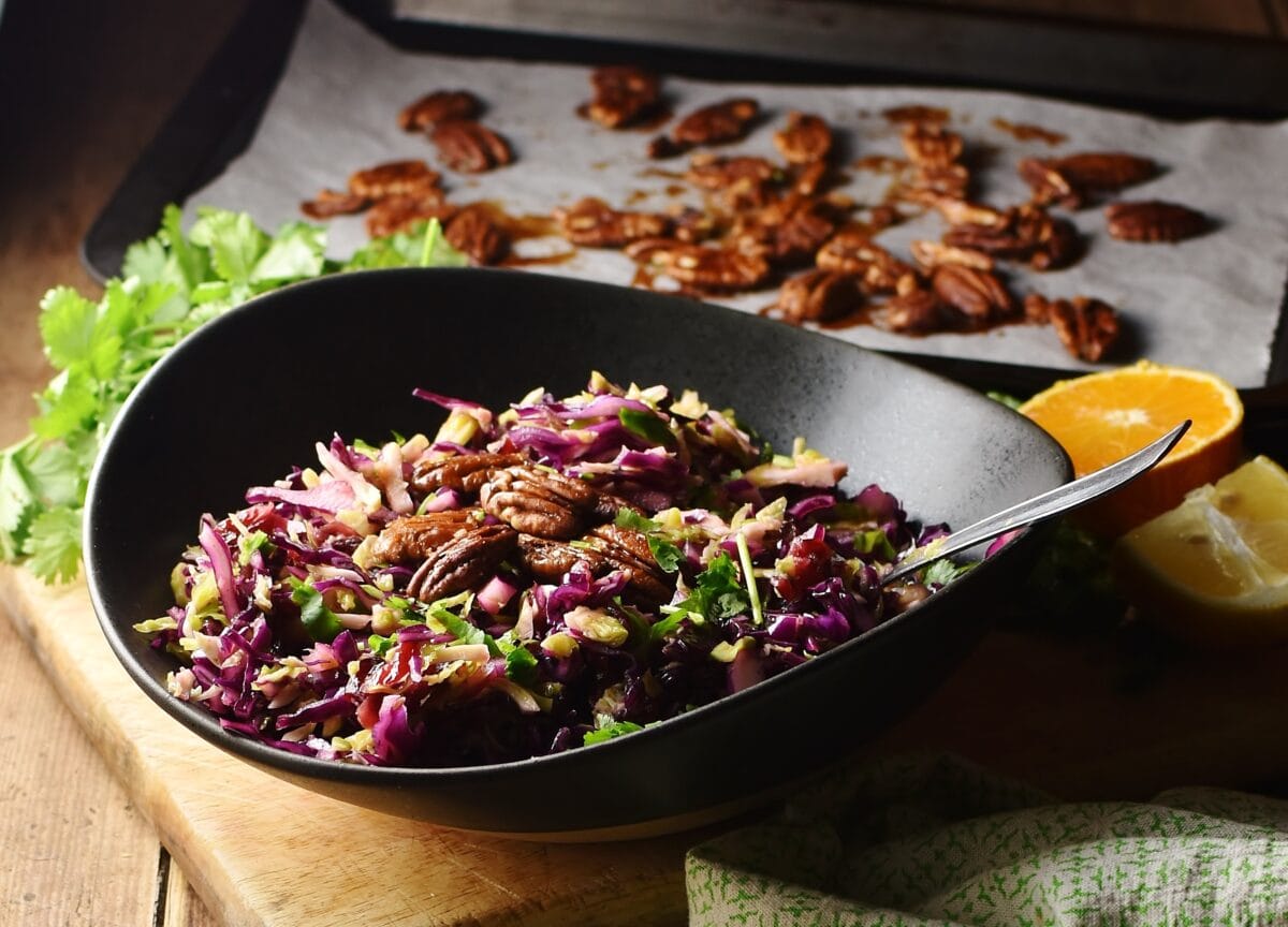 Red cabbage Christmas slaw with pecans and herbs in black bowl with spoon, and pecans on top of baking sheet with parchment paper in background.