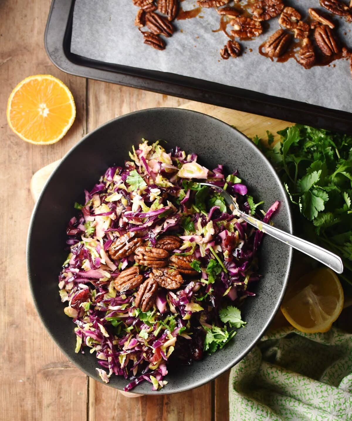 Shredded red cabbage, brussels sprouts, dried cranberries, pecans and herbs in oval black bowl with spoon, herbs, orange half and pecans on top of baking sheet in background.