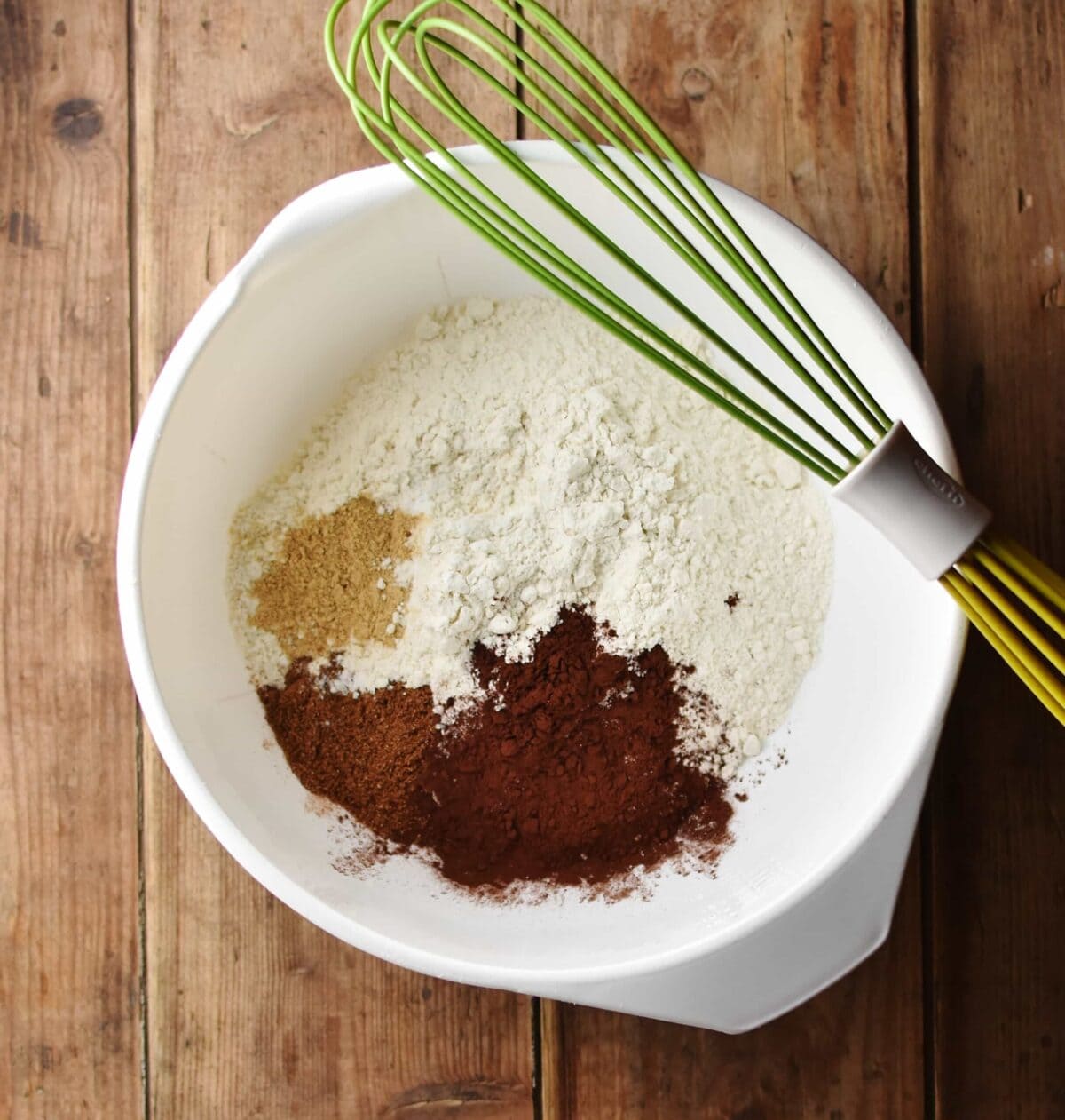 Flour, cocoa and spices in large white bowl with green whisk.