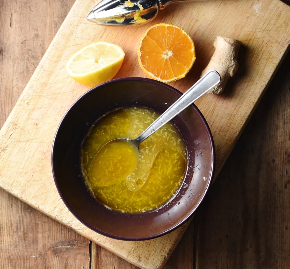 Orange marinade mixture in purple bowl with spoon, orange, lemon and ginger in background, on top of wooden board.