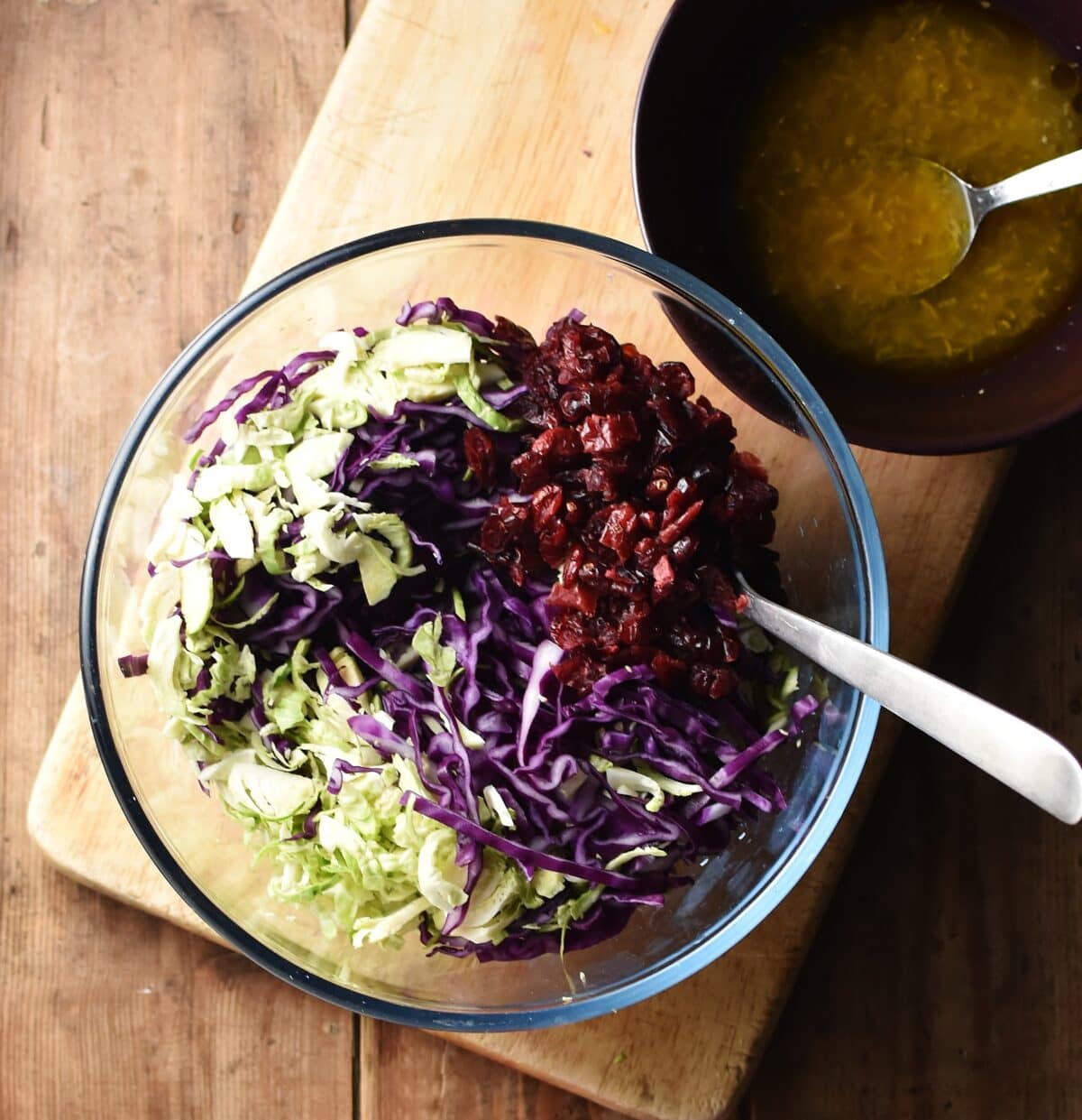Shredded red cabbage, brussels sprouts and dried cranberries in mixing bowl with spoon and marinade mixture in bowl in background.