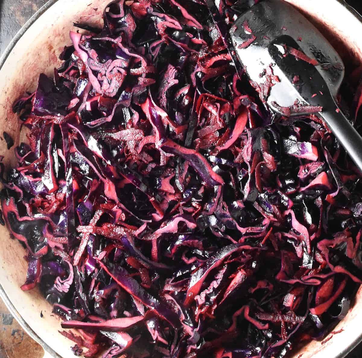 Close-up view of cooking shredded red cabbage in large shallow pan with black spoon.