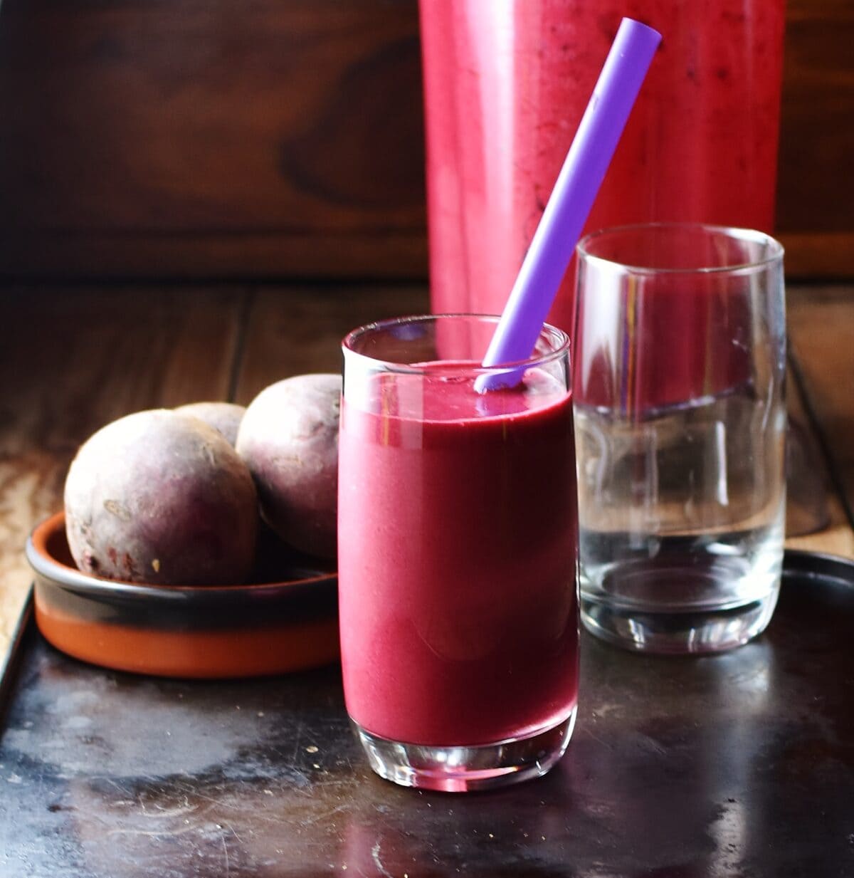 Side view of beetroot smoothie with purple straw in glass, with empty glass, beetroot in brown dish and blender in background.