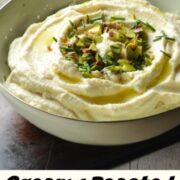 Close-up view of cauliflower dip garnished with nuts and herbs inside green bowl with wooden board and herbs in background.